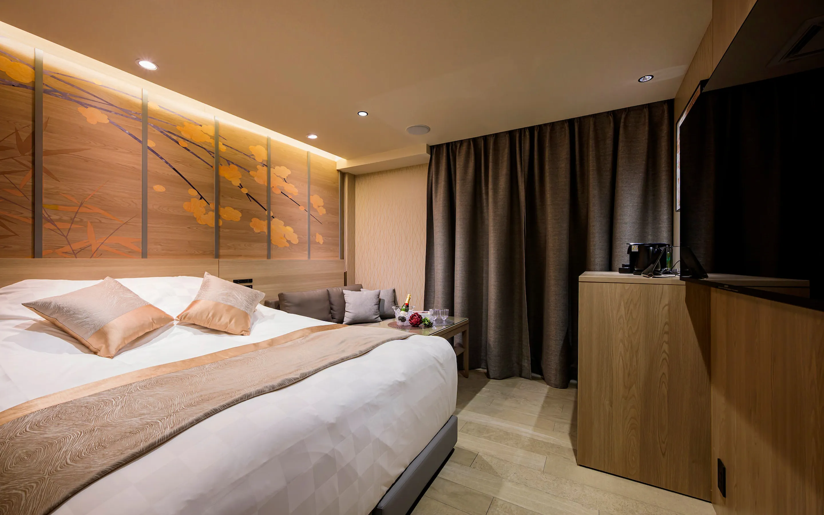 モダンで落ち着いた和洋室のベッドルーム。壁面に木が描かれた木製パネルとシャンパンが置かれたリビングスペース。