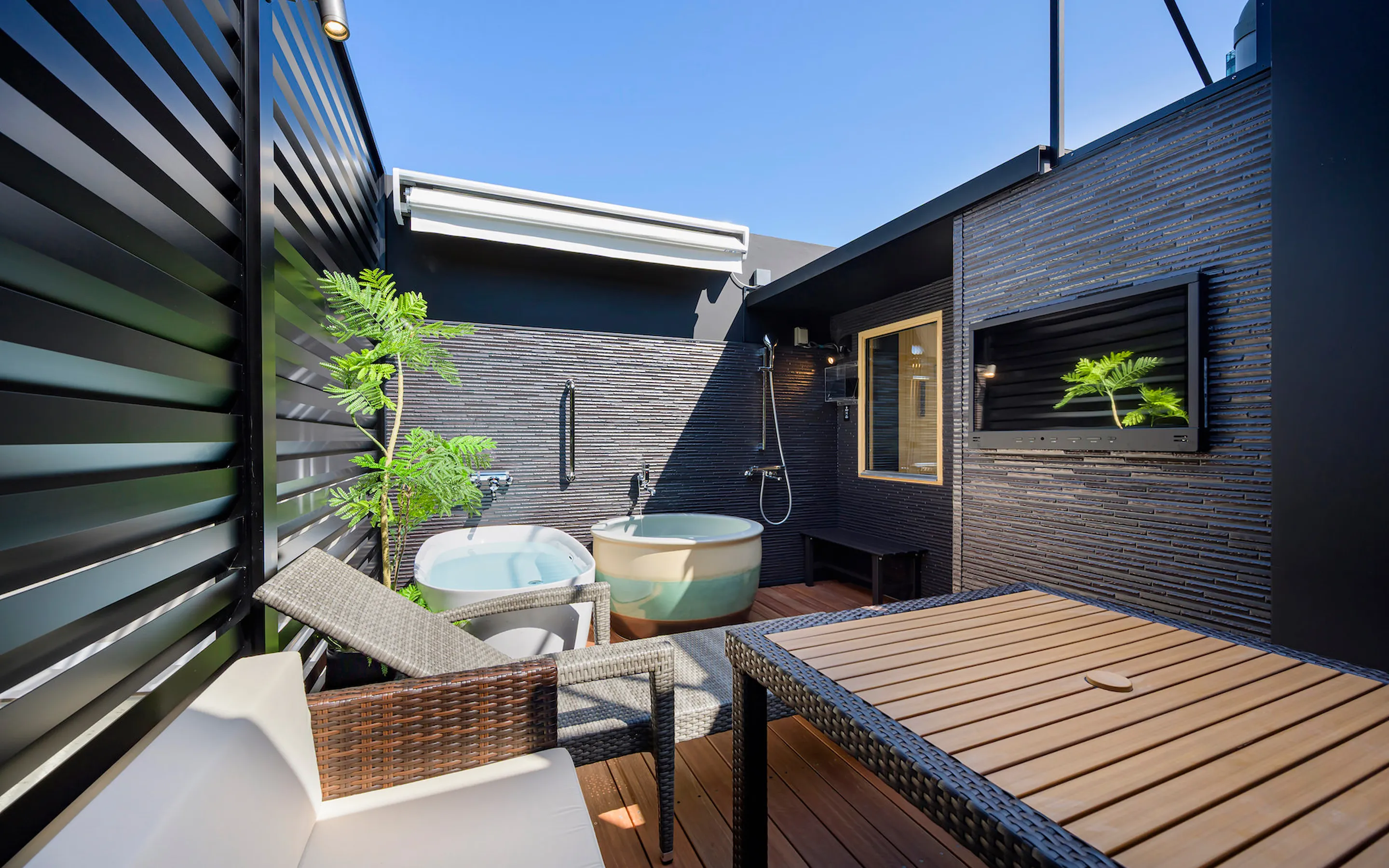 屋外テラスにある2つの露天風呂。ウッドデッキにはラタン調のソファとテーブル、屋外用テレビが設置された開放的な空間。