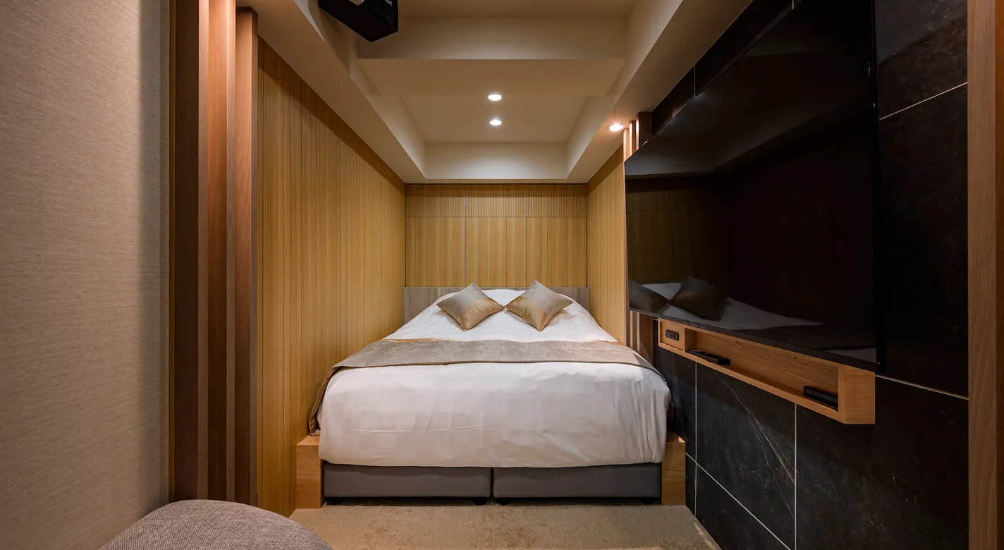 ベッド正面に大型モニターを設置したシアタールーム仕様の客室。木目調の壁に囲まれたコンパクトで没入感のある設計