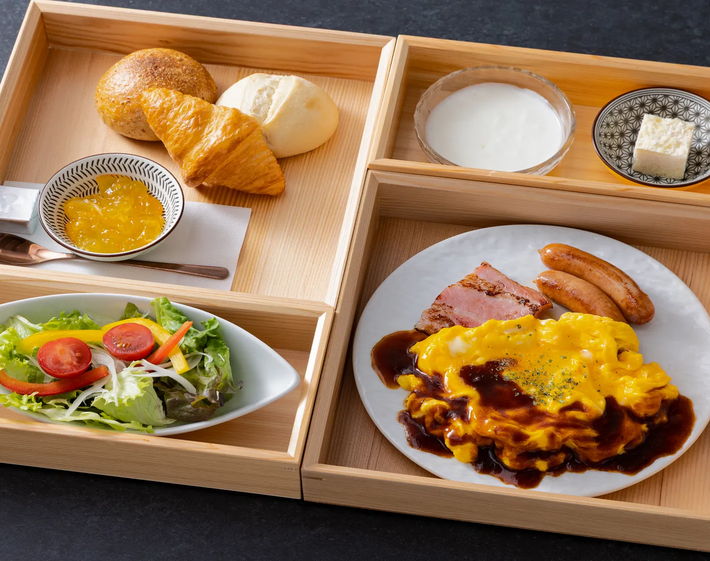木製トレイに盛り付けられた朝食セット。デミグラスソースのオムレツ、ベーコン、ソーセージ、焼きたてパン、サラダ、ヨーグルト